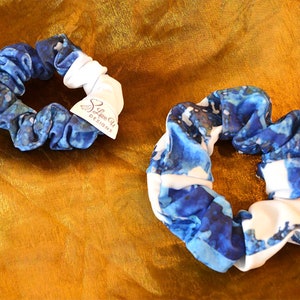 Peut inclure: Deux chouchous bleus et blancs avec un motif ressemblant à une aquarelle. Les chouchous sont en tissu et ont un élastique blanc. Le plus grand chouchou a une étiquette blanche avec le texte "Love Chi Designs".