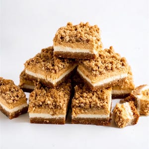 Peut inclure: Une pile de barres dessert carr&eacute;es &agrave; l'aspect multicouche. Elles pr&eacute;sentent une base marron fonc&eacute;, une couche blanche et un crumble brun clair. Les barres sont dispos&eacute;es sur une surface blanche, mettant en valeur leurs textures.