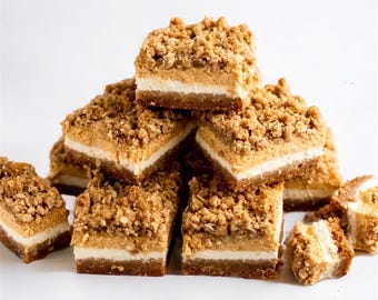 Puede incluir: Una pila de barras de postre cuadradas con una apariencia en capas. Tienen una base marrón oscuro, una capa blanca y una cobertura de migas de color marrón claro. Las barras están dispuestas sobre una superficie blanca, mostrando sus texturas.
