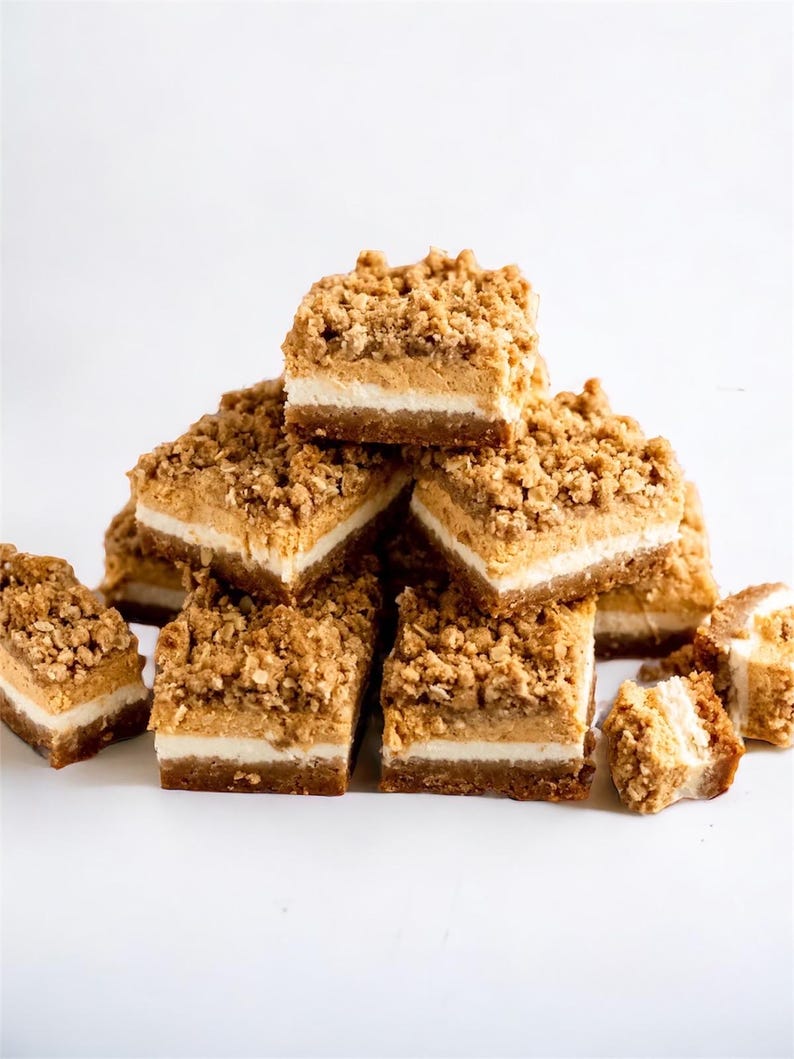 Peut inclure: Une pile de barres dessert carr&eacute;es &agrave; l'aspect multicouche. Elles pr&eacute;sentent une base marron fonc&eacute;, une couche blanche et un crumble brun clair. Les barres sont dispos&eacute;es sur une surface blanche, mettant en valeur leurs textures.