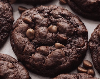 Puede incluir: Primer plano de varias galletas de chocolate recién horneadas con chispas de chocolate. Las galletas son de un color marrón oscuro intenso y tienen una superficie ligeramente texturizada. Algunas chispas de chocolate son visibles encima de las galletas.