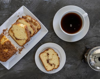 Puede incluir: Vista aérea de pan de canela en rodajas sobre un plato rectangular blanco, con una taza de café en un platillo y una sola rebanada en un plato pequeño. También se ve una prensa francesa. La comida está sobre una superficie gris oscura.