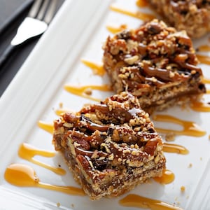 Puede incluir: Tres barras de caramelo y nuez pecana en un plato blanco con salsa de caramelo rociada sobre ellas. Las barras est&aacute;n hechas con una corteza de galleta, una capa de caramelo y una cobertura de nueces pecanas.