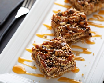 Puede incluir: Tres barras de caramelo y nuez pecana en un plato blanco con salsa de caramelo rociada sobre ellas. Las barras están hechas con una corteza de galleta, una capa de caramelo y una cobertura de nueces pecanas.