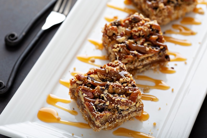 Puede incluir: Tres barras de caramelo y nuez pecana en un plato blanco con salsa de caramelo rociada sobre ellas. Las barras est&aacute;n hechas con una corteza de galleta, una capa de caramelo y una cobertura de nueces pecanas.