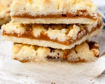 Puede incluir: Una pila de tres barras de shortbread con caramelo. Las barras tienen una cobertura desmenuzable de color marrón dorado y una gruesa capa de relleno de caramelo. Están apiladas sobre un plato blanco con diseño moteado, sobre fondo blanco.