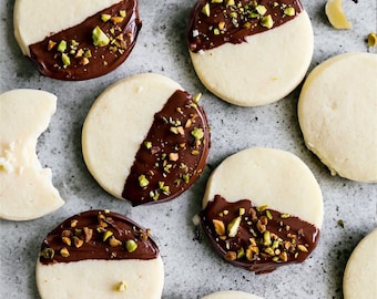 Puede incluir: Vista aérea de varias galletas de mantequilla redondas, sumergidas a la mitad en chocolate negro y espolvoreadas con pistachos picados. Algunas galletas están enteras, mientras que otras han sido parcialmente comidas. Las galletas están dispuestas sobre una superficie gris claro.