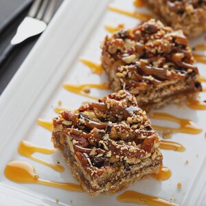 Puede incluir: Tres cuadrados de barras de caramelo y nuez pecana en un plato rectangular blanco con un chorrito de salsa de caramelo. Las barras est&aacute;n cubiertas con nueces pecanas y una capa de caramelo.