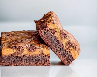 Puede incluir: Dos brownies recién horneados con aspecto veteado. Los brownies tienen una base de chocolate negro con un remolino más claro, color caramelo en la parte superior. La imagen es un primer plano, que destaca la textura y las capas del postre.