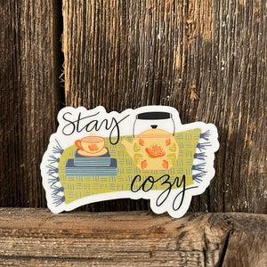 May include: A white sticker with a green and blue patterned rug, a teapot and a teacup, and the text "Stay cozy".