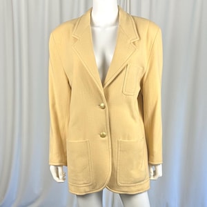 Puede incluir: Un blazer amarillo claro con dos botones dorados y dos bolsillos delanteros. El blazer se exhibe en un maniquí. El blazer tiene un diseño clásico y está hecho de un material suave. El blazer es una talla mediana.
