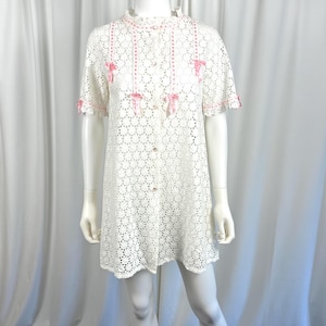 Könnte beinhalten: Weißes Lochmusterkleid mit kurzen Ärmeln und Knopfleiste. Das Kleid hat rosa Schleifen und Bänder. Das Kleid wird auf einer Schaufensterpuppe präsentiert.