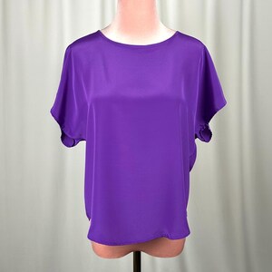 Puede incluir: Una blusa de color morado vibrante con mangas cortas, acabado suave y satinado, escote redondo sencillo y una silueta relajada y fluida. La prenda presenta un dobladillo recto y mangas holgadas que caen de forma natural sobre los hombros.