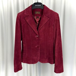 May include: A burgundy suede blazer with a classic notched collar and two-button closure. The jacket features long sleeves and a tailored fit, hanging on a black hanger. The brand name "DANIER" is visible on the hanger.