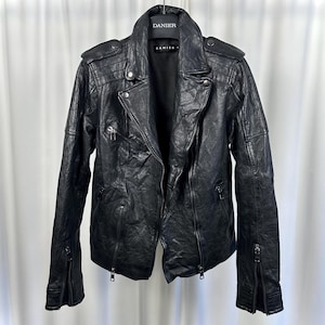 May include: Black leather jacket with an asymmetrical zip, multiple zipped pockets, and a notched collar. The jacket has a textured finish and is hanging on a hanger. The brand name "DANIER" is visible on the hanger.