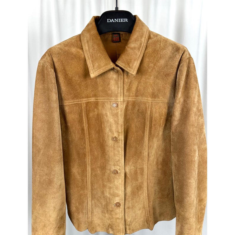 May include: A tan suede jacket with a collared neckline and button-down closure. The jacket features visible stitching details and is hanging on a black hanger. The brand name "DANIER" is visible on the hanger.