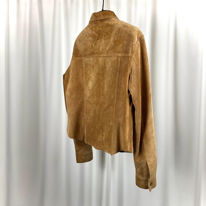May include: A tan suede jacket with a buttoned cuff and a collared neckline. The jacket is hanging on a black hanger against a white backdrop. The back of the jacket is visible, showcasing the seams and the texture of the suede.