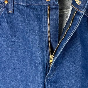 May include: Close-up of a pair of blue denim jeans with a brass zipper and a button closure. The zipper is partially open, revealing the inside of the jeans.