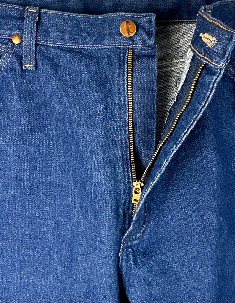 May include: Close-up of a pair of blue denim jeans with a brass zipper and a button closure. The zipper is partially open, revealing the inside of the jeans.