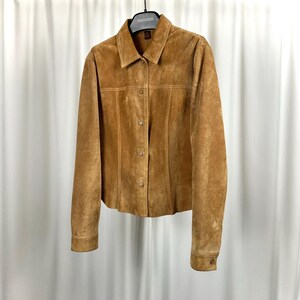 May include: A tan suede jacket with a collared neckline and button-down closure. The jacket has long sleeves with buttoned cuffs and vertical seam detailing. It is hanging on a black hanger against a white backdrop.