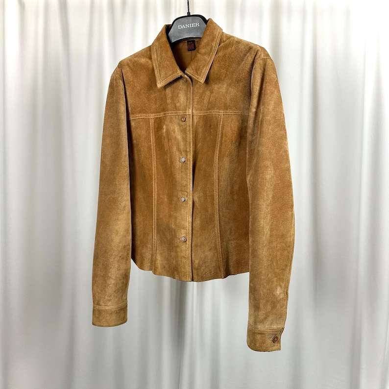 May include: A tan suede jacket with a collared neckline and button-down closure. The jacket has long sleeves with buttoned cuffs and vertical seam detailing. It is hanging on a black hanger against a white backdrop.