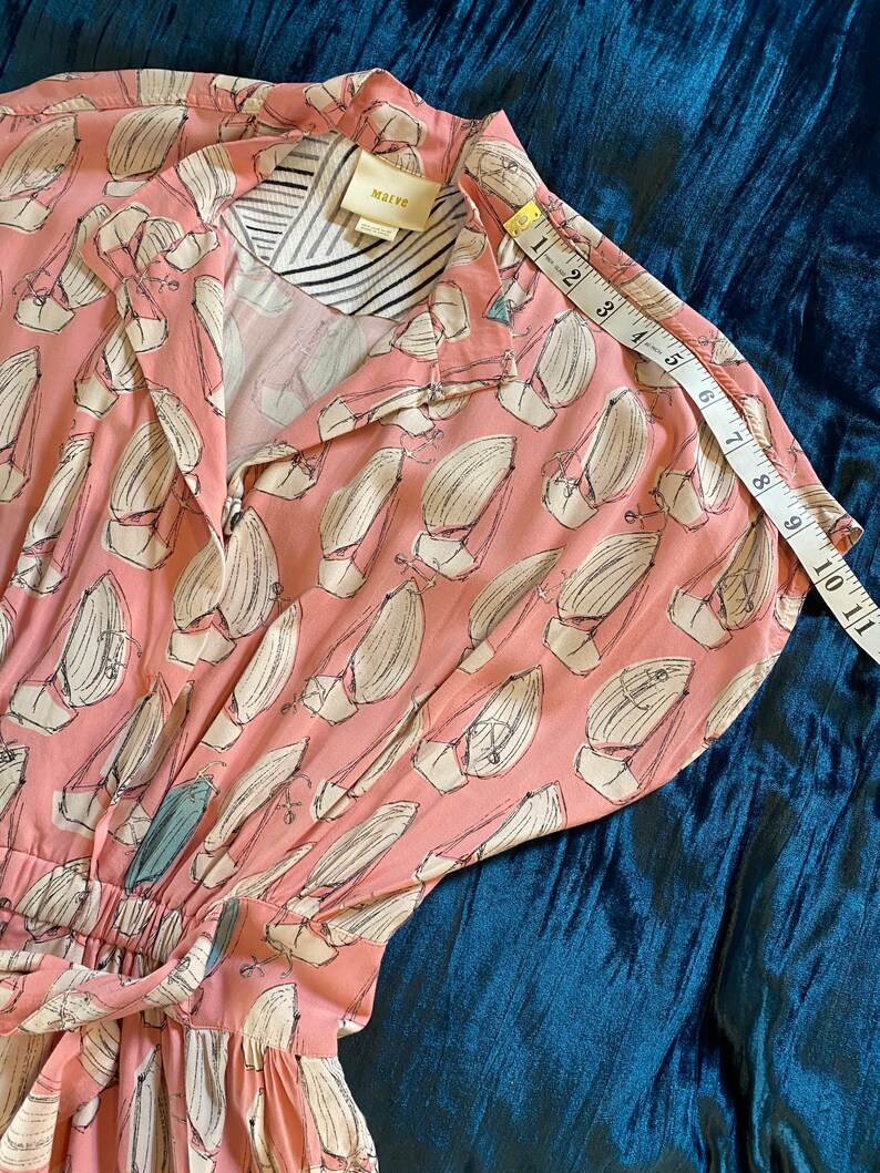May include: A pink and white dress with a sailboat print. The dress has a button-up front and a tie at the waist. The dress is made of a lightweight fabric and has short sleeves. The dress is labeled "Malva" and has a size tag.