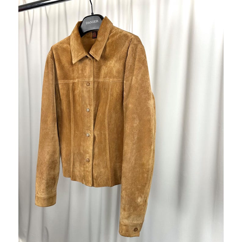 May include: A tan suede jacket with a collared neckline and button-up front. The jacket has long sleeves and a tailored fit, hanging on a black hanger. The jacket is a neutral colour and appears to be made of soft suede material.