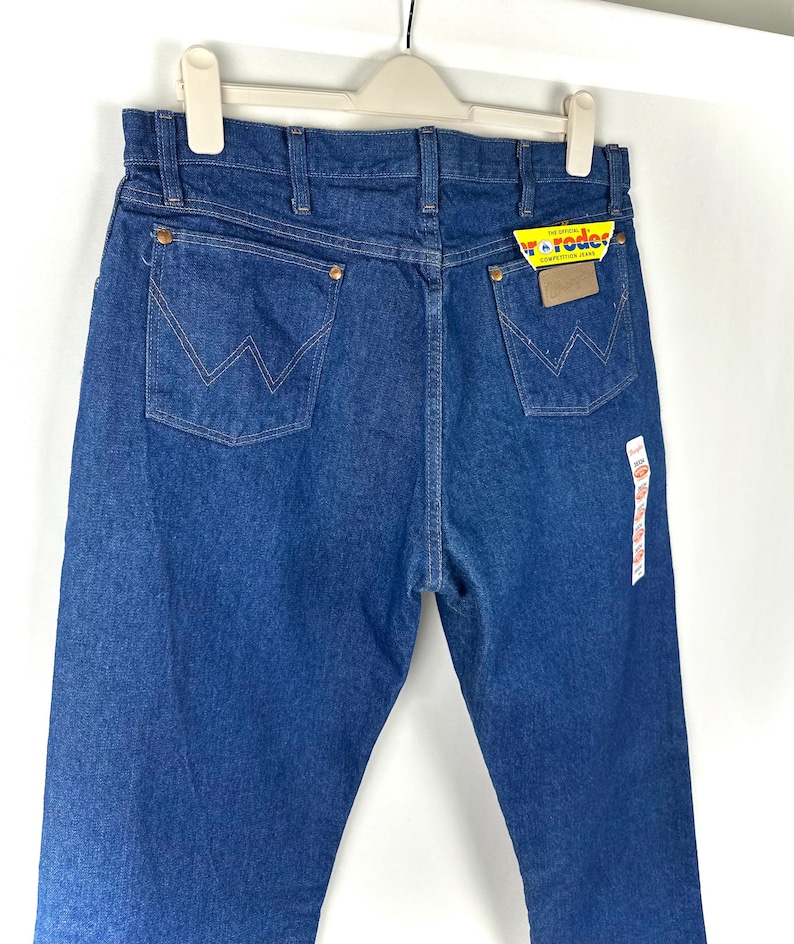 May include: A pair of blue denim jeans with a yellow and red Wrangler label attached. The jeans have a classic five-pocket style and a straight leg.