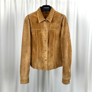 May include: A tan suede jacket with a button-up front and long sleeves. The jacket has a collared neckline and is hanging on a black hanger. The brand name "DANIER" is visible on the hanger.