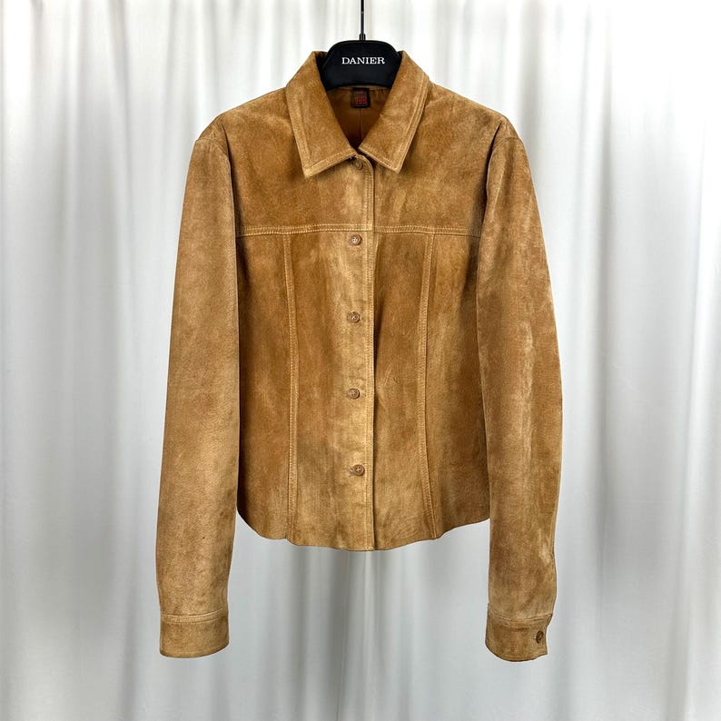 May include: A tan suede jacket with a button-up front and long sleeves. The jacket has a collared neckline and is hanging on a black hanger. The brand name "DANIER" is visible on the hanger.