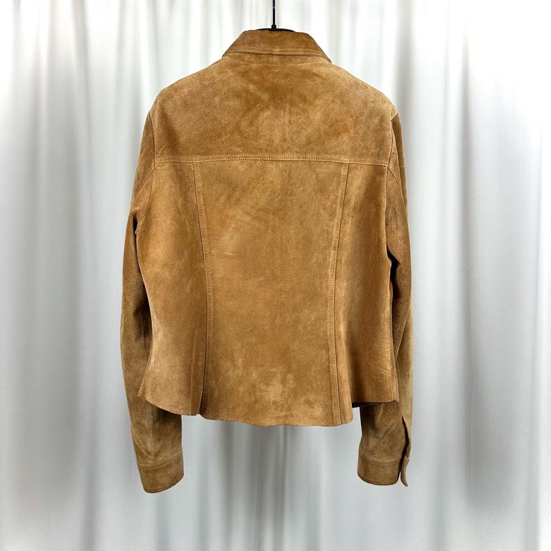 May include: A tan suede jacket with a button-down front, long sleeves, and a collar. The back of the jacket is visible, showing the seams and the texture of the suede. The jacket is hanging on a black hanger against a white backdrop.