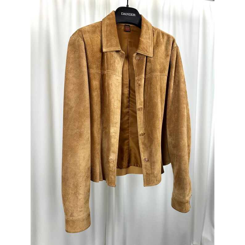 May include: A tan suede jacket with a button-up front and collared neckline. The jacket is hanging on a black hanger against a white backdrop. The jacket has long sleeves and a tailored fit, suitable for outerwear.