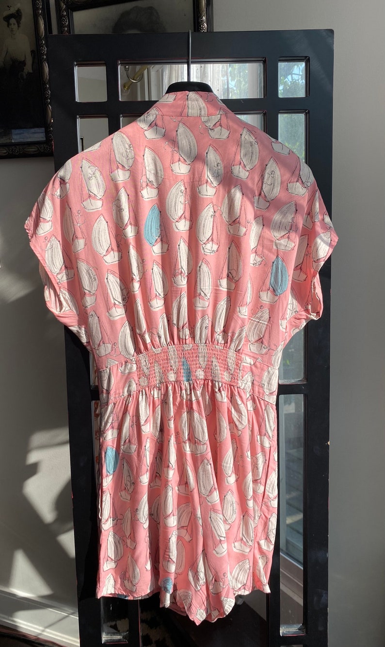 May include: A pink and white short-sleeved jumpsuit with a sailboat print. The jumpsuit has an elastic waistband and a relaxed fit.