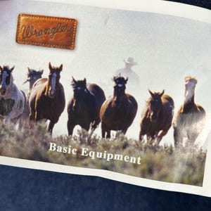 May include: A vintage Wrangler advertisement featuring a group of horses running through a field. The text "Basic Equipment" is printed below the image. The Wrangler logo is in the top left corner.