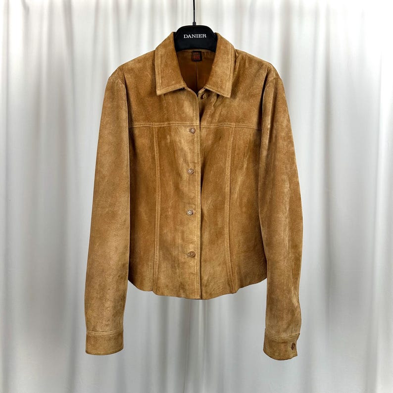 May include: A tan suede jacket with a button-up front and a collared neckline. The jacket has long sleeves and is hanging on a black hanger. The brand name "DANIER" is visible on the hanger.