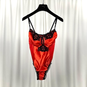 May include: A red satin bodysuit with black lace trim and accents. The lingerie features adjustable straps and a keyhole detail at the bust. The garment is hanging on a black hanger against a white backdrop.