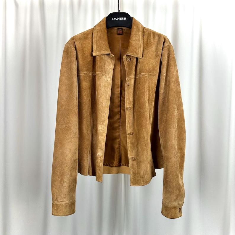 May include: A tan suede jacket with a collared neckline and button-up closure. The jacket is hanging on a black hanger with the brand name "DANIER" printed on it. The jacket has long sleeves and a classic, tailored design.