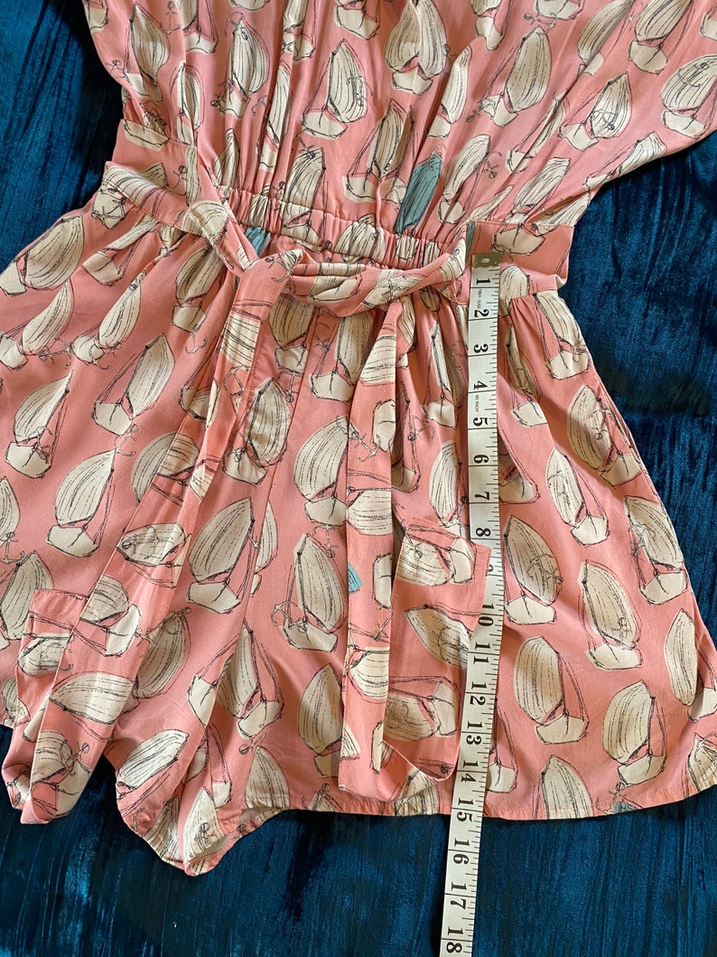 May include: A pink and white patterned short-sleeved romper with a tie waist. The romper has a sailboat print and is measured with a tape measure.