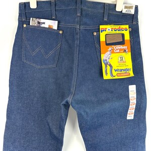 May include: A pair of blue denim jeans with a Wrangler label. The jeans are a cowboy cut and have a 'Pro Rodeo' tag attached. The tag says 'The Official Competition Jeans' and 'Cowboy Cut 13 oz.'