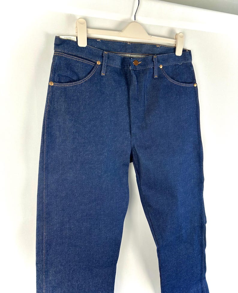 May include: A pair of dark blue denim jeans hanging on a white hanger. The jeans have a classic straight leg style and are made of a sturdy, durable fabric.
