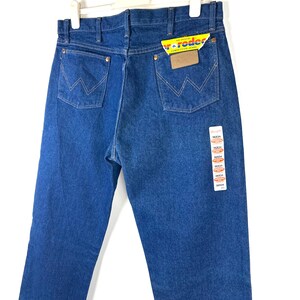 May include: A pair of blue denim jeans with a brown leather patch on the back pocket. The jeans have a tag attached with the text "Wrangler" and "The Original Competition Jeans".