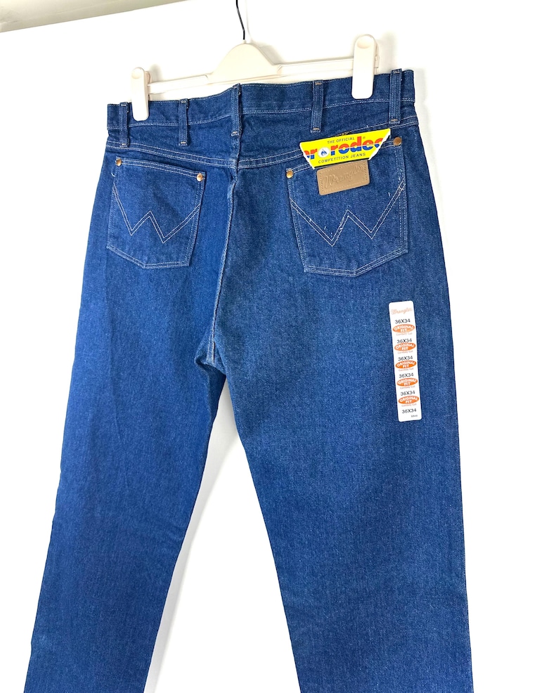May include: A pair of blue denim jeans with a brown leather patch on the back pocket. The jeans have a tag attached with the text "Wrangler" and "The Original Competition Jeans".