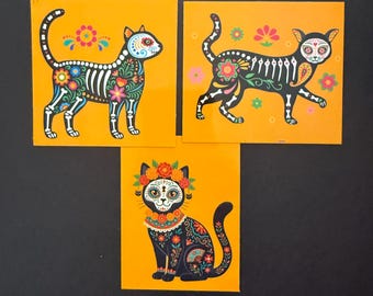 May include: Three square prints on a bright orange background featuring black cats decorated with colorful floral patterns and skeletal motifs. Two cats are shown in profile walking, while the third cat is sitting and wearing a floral crown.