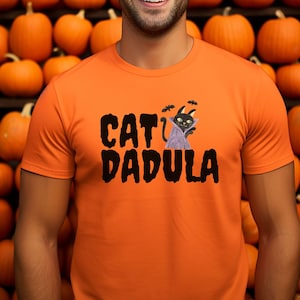 May include: Orange t-shirt with the text "CAT DADULA" in black, featuring a cartoon cat dressed as a vampire with bats. The shirt is a Halloween-themed design, perfect for cat lovers and spooky season.