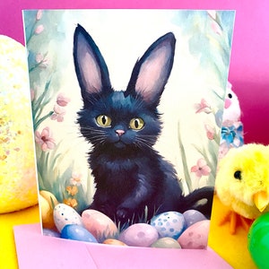 May include: A greeting card featuring a black cat with long ears, resembling a rabbit, surrounded by Easter eggs. The illustration has a watercolor style with soft colors and floral accents, set against a pink background.