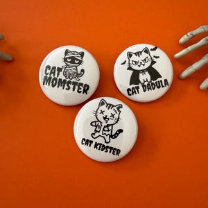 May include: Three round white pinback buttons featuring black graphic illustrations of cats in Halloween costumes. One button shows a mummy cat labeled CAT MOMSTER, one shows a vampire cat labeled CAT DADULA, and one shows a zombie cat labeled CAT KIDSTER.