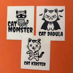 May include: Three square stickers featuring black line art of cats on white backgrounds. One shows a mummy cat labeled CAT MOMSTER, one a vampire cat labeled CAT DADULA with bats, and one a zombie cat labeled CAT KIDSTER with stitches and drool.