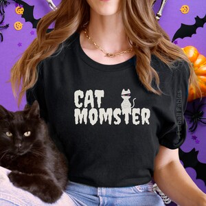 May include: Black t-shirt with the text "CAT MOMSTER" in white, featuring a mummy cat graphic. A black cat is in the foreground. The shirt is part of a Halloween-themed collection.