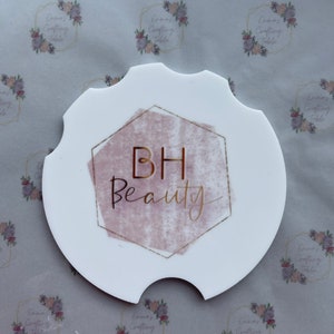 May include: White car coaster with a unique shape, featuring the text "BH Beauty" in gold script within a pink and gold geometric design. The coaster has a cutout for easy removal and placement in a car cup holder.