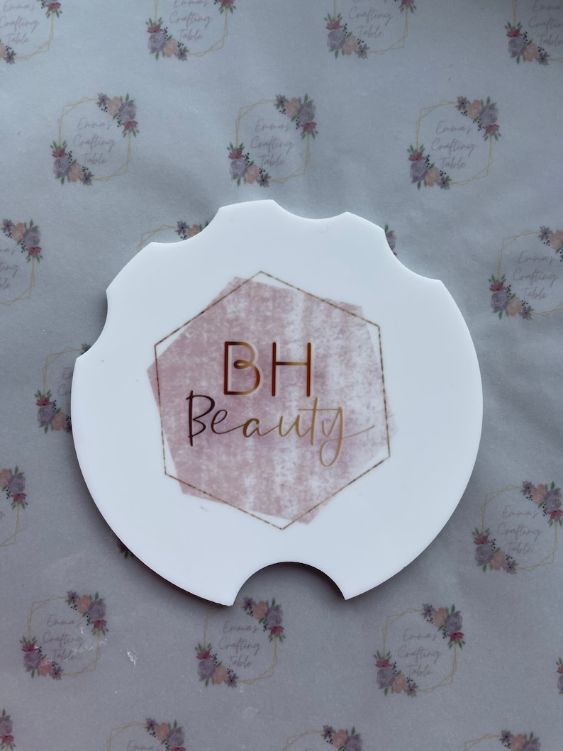May include: White car coaster with a unique shape, featuring the text "BH Beauty" in gold script within a pink and gold geometric design. The coaster has a cutout for easy removal and placement in a car cup holder.